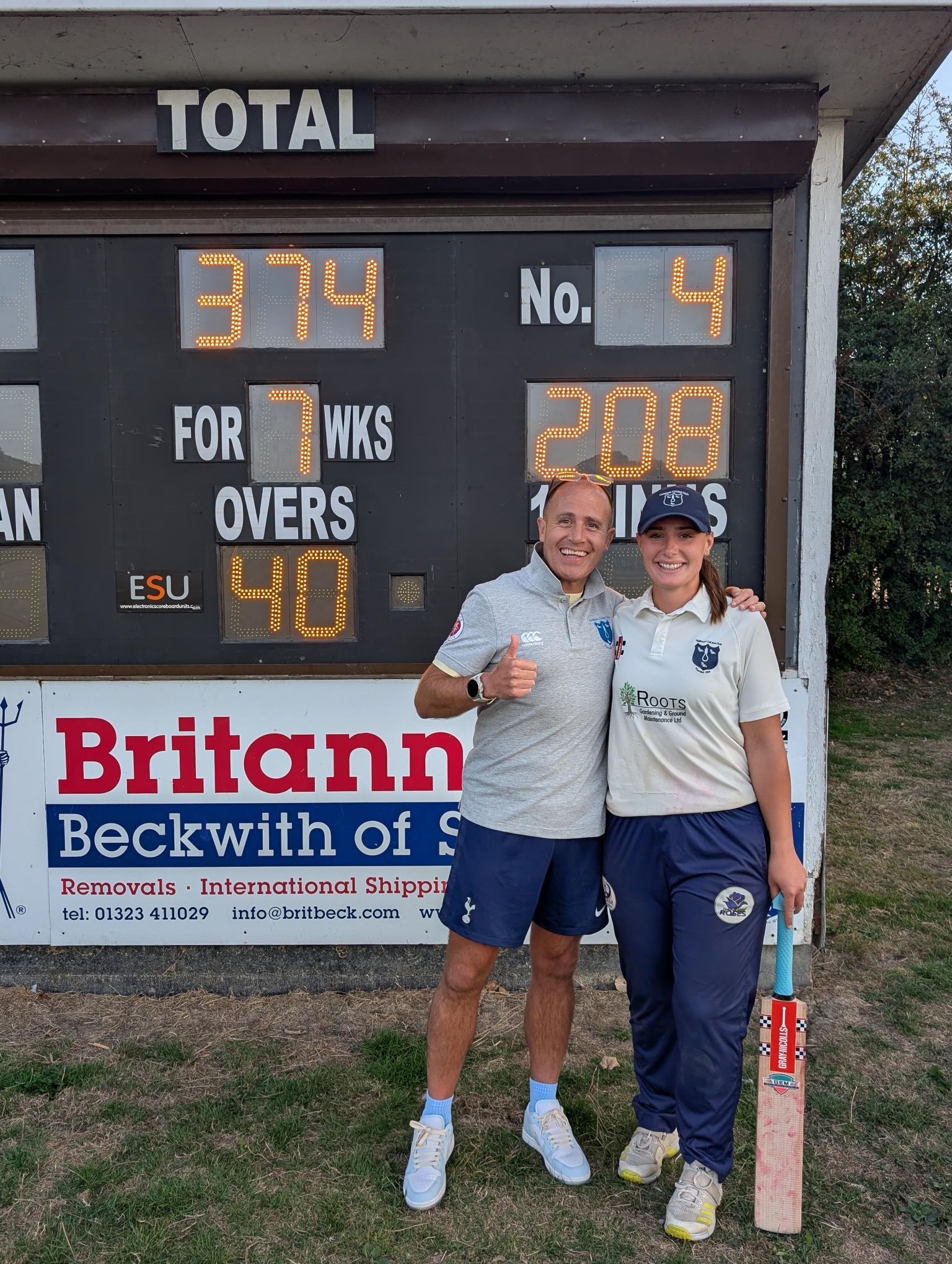 Hollie and Ben standing in front of the scoreboard displaying a total of 374 for 7, with the number 4 batter (Hollie) having scored 208 runs during the innings.
