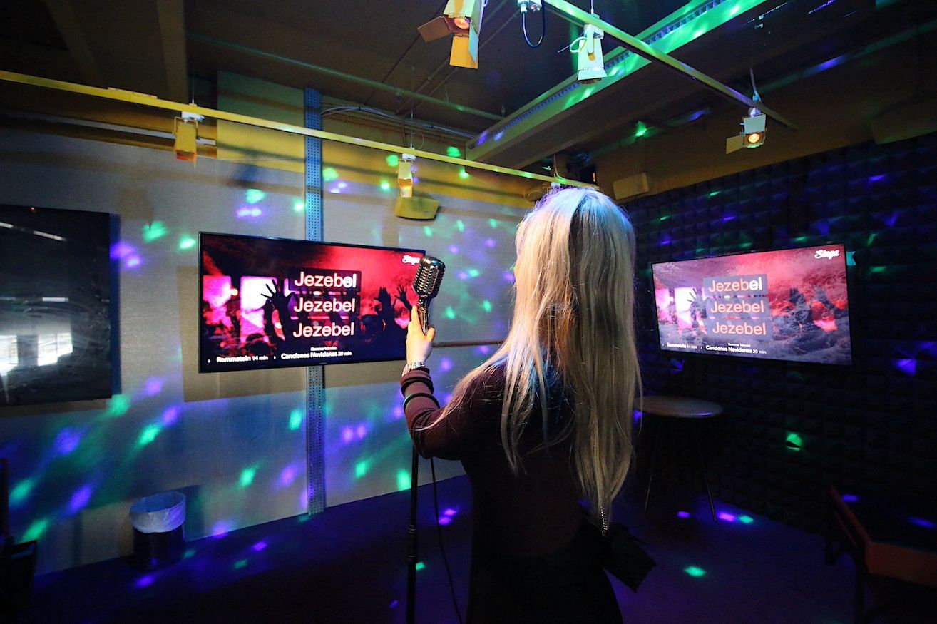 Kvinna sjunger karaoke i ett rum med två skärmar och neonljus.