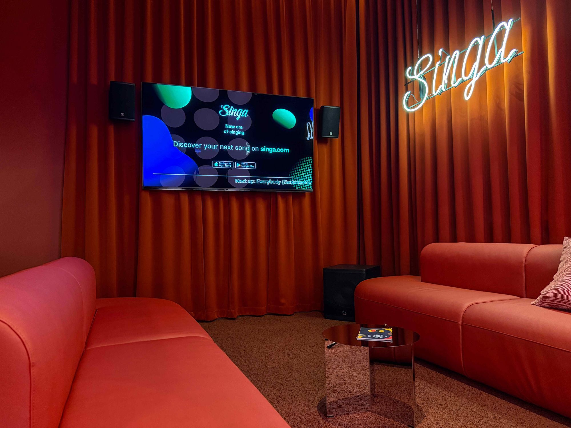 Karaoke-Raum mit Bildschirm, Lautsprechern und roten Sofas.
