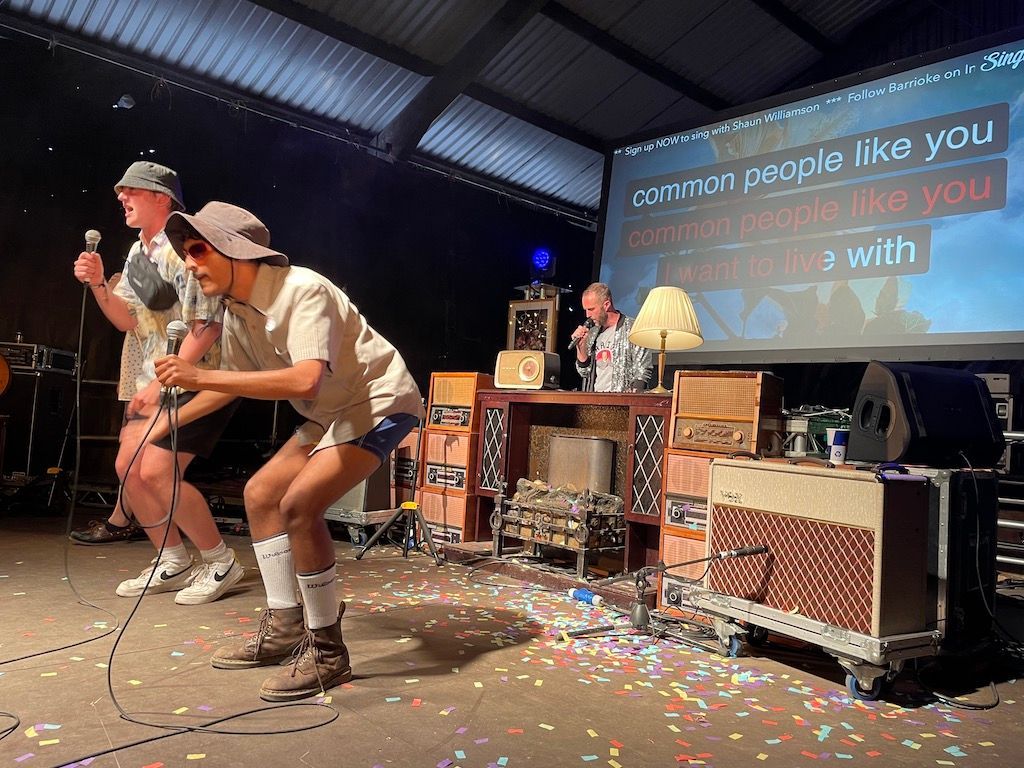 Moderiertes Karaoke: Zwei junge Männer performen Karaoke auf einer Bühne vor dem Karaoke-Moderator.