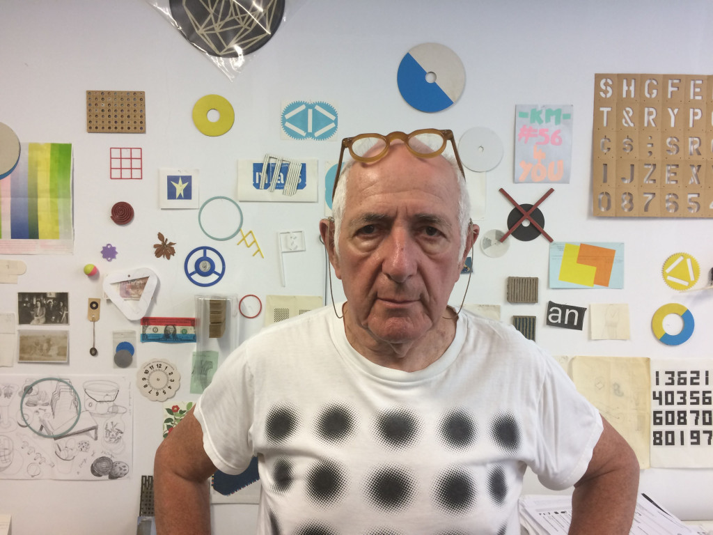 Karel Martens, portret in zijn studio, met grafische ontwerpen op de achtergond – speels en kleurrijk. Foto door zijn zoon Diederik