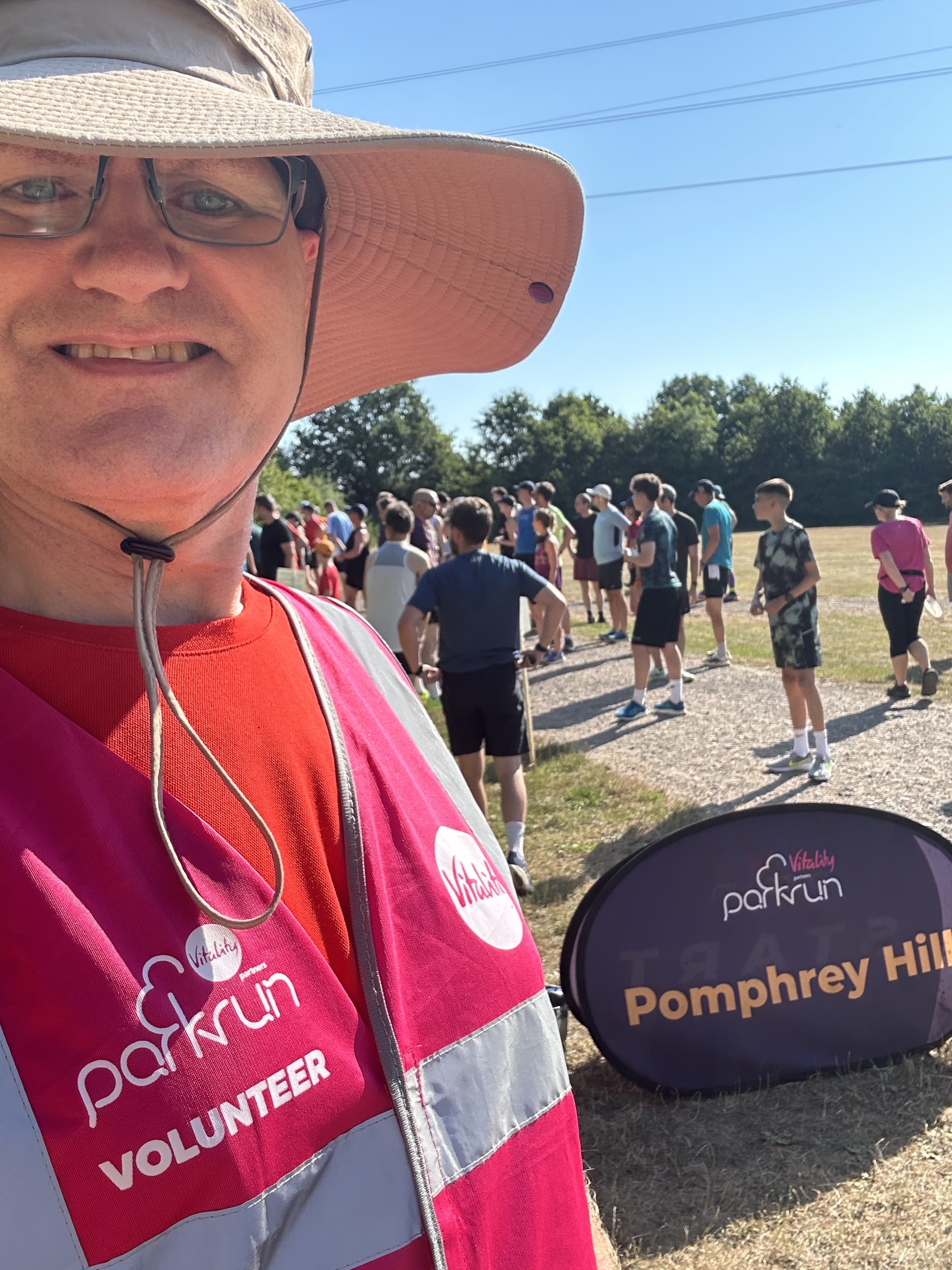 Selfie of Richard in front of the Pomphrey Hill parkrun sign. I’m wearing a very wide brimmed sun hat, and a pink florescent parkrun volunteer vest. Runners in the background are getting ready for the start. It’s sunny and very hot.