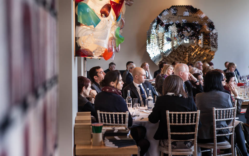 Group of people gathered around round tables in a room displaying contemporary art concentrating on a speaker who is out of the frame.