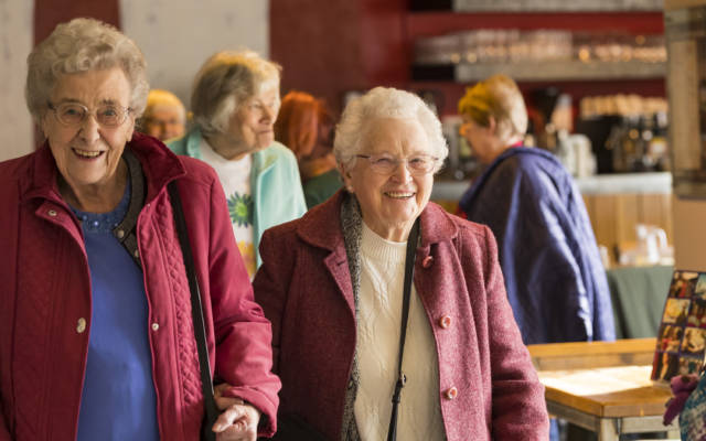 Two smiling older ladies in coats walking through cafe with linked arms