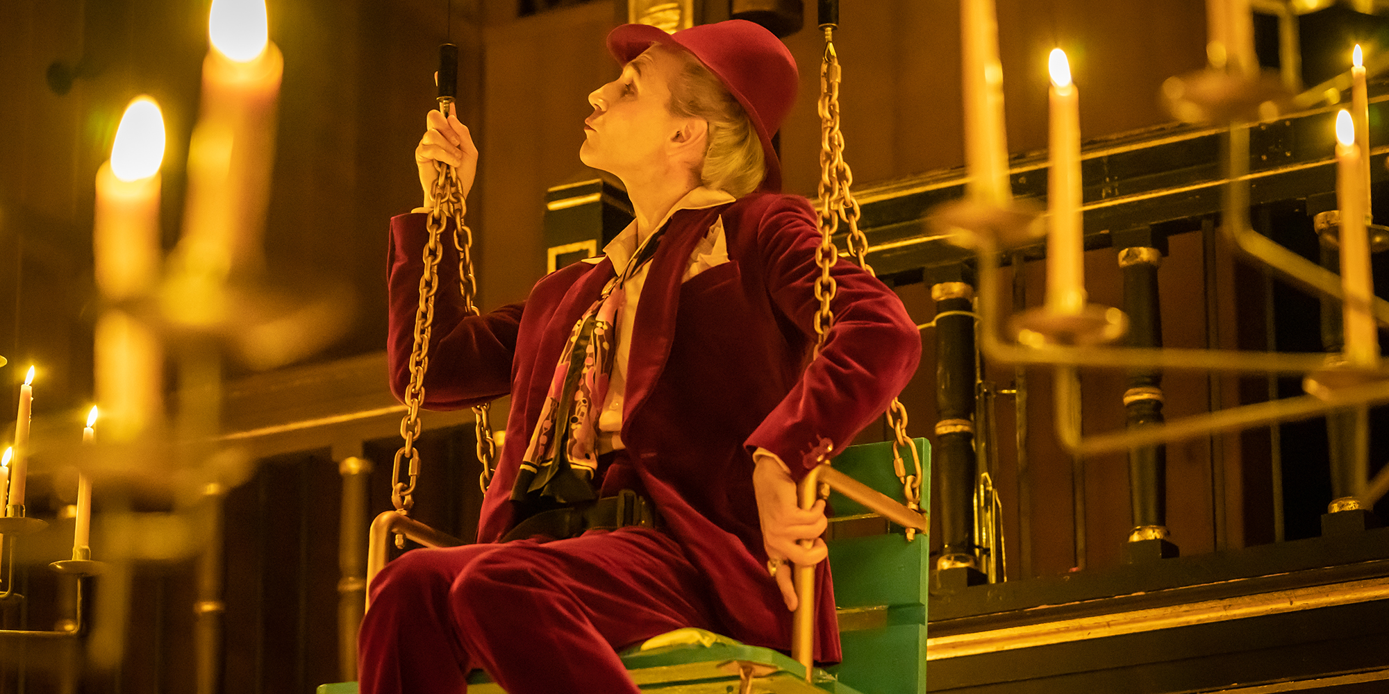 After Edward Such Stuff Shakespeare's Globe An actor wearing a deep red velvet suit sits on a green swing surrounded by lit candles.