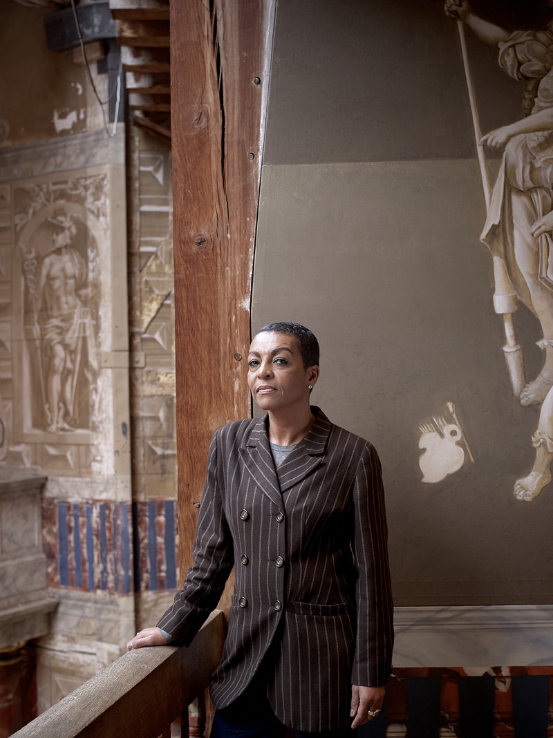 Adjoa Andoh A black woman with short cropped hair stands against a grey wall wearing a dark pinstripe suite.