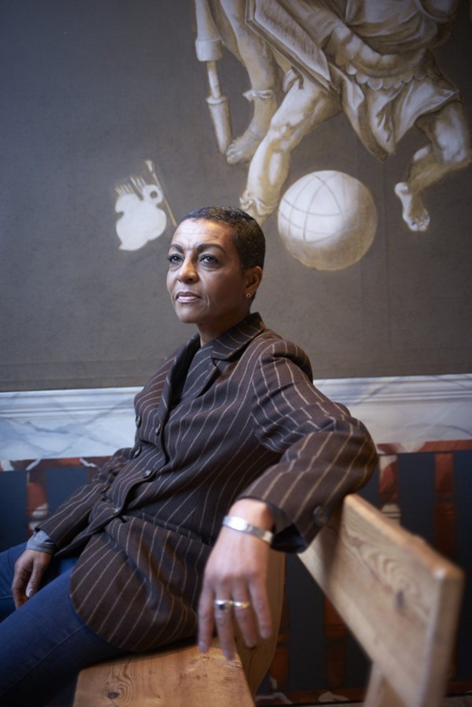 Adjoa-Andoh-3 A black woman with short cropped hair stands against a grey wall wearing a dark pinstripe suite.