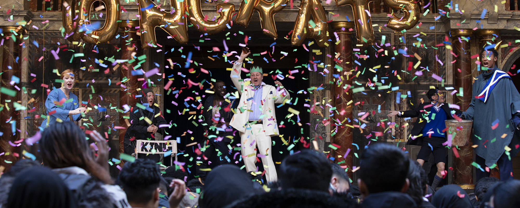 Playing Shakespeare Macbeth 2020 webite masthead Actors on stage dancing at a party and confetti falling all around them