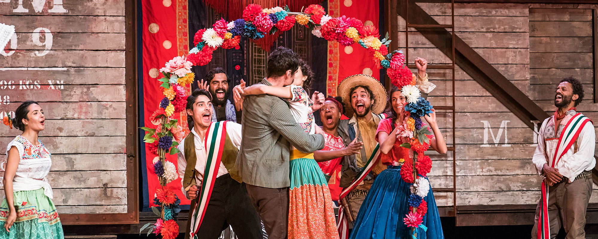 Shakespeare Diaries_Much Ado About Nothing 2017 _ Such Stuff s5 e4_ masthead Beatrice and Benedick embrace and kiss surrounded by onlookers. The stage is full of brightly coloured decorations and costumes.