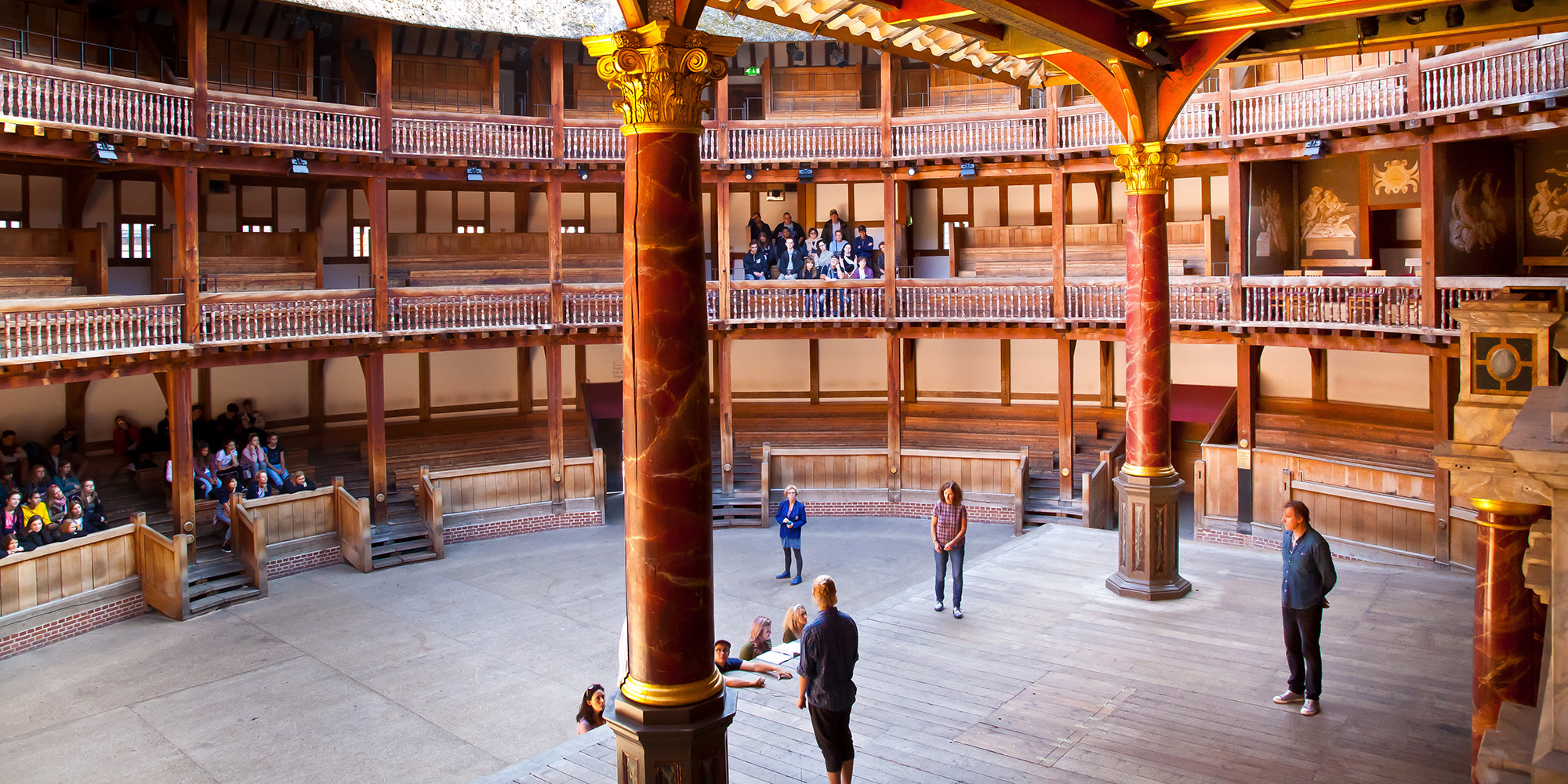 Freelance task force Shakespeare's Globe People stand on a wooden thrust stage of a circular timbre structured theatre.