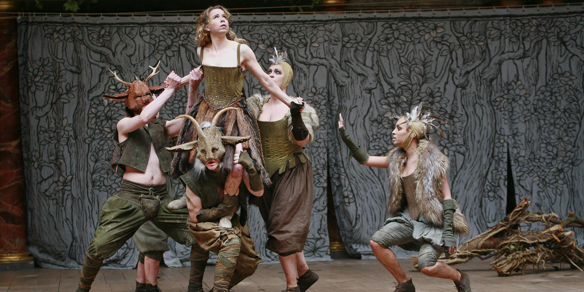 Midsummer's Day YouTube Dream Shakespeare's Globe A woman wearing a green corset dress is lifted into the air by a group of people wearing fur and antlers.
