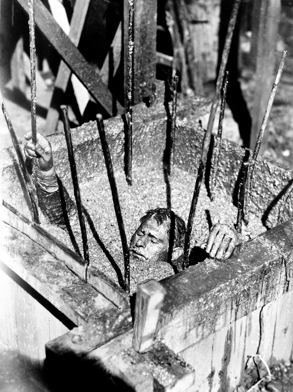 Sam Wanamaker buried in liquid concrete for the filming of Give Us This Day. A man is buried to his neck in gravel, behind bars.
