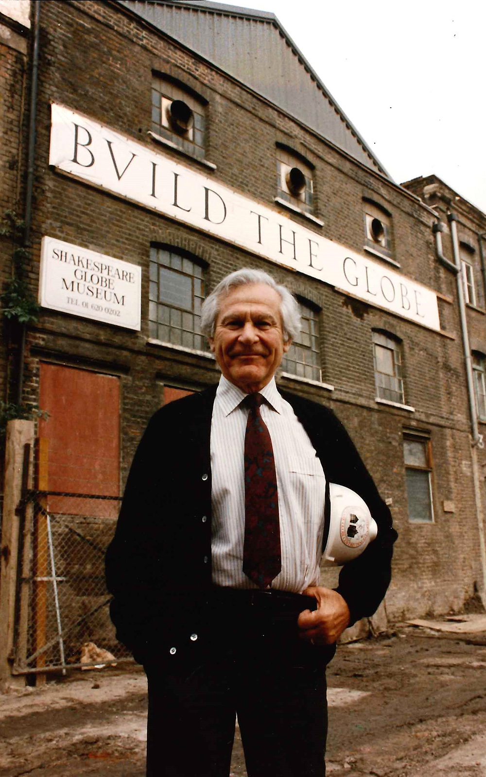 Sam Wanamaker Globe Museum Shakespeare's Globe A man in a suit and holding a hardhat stands before a warehouse building with the sign reading: Rebuilding Shakespeare's Globe.