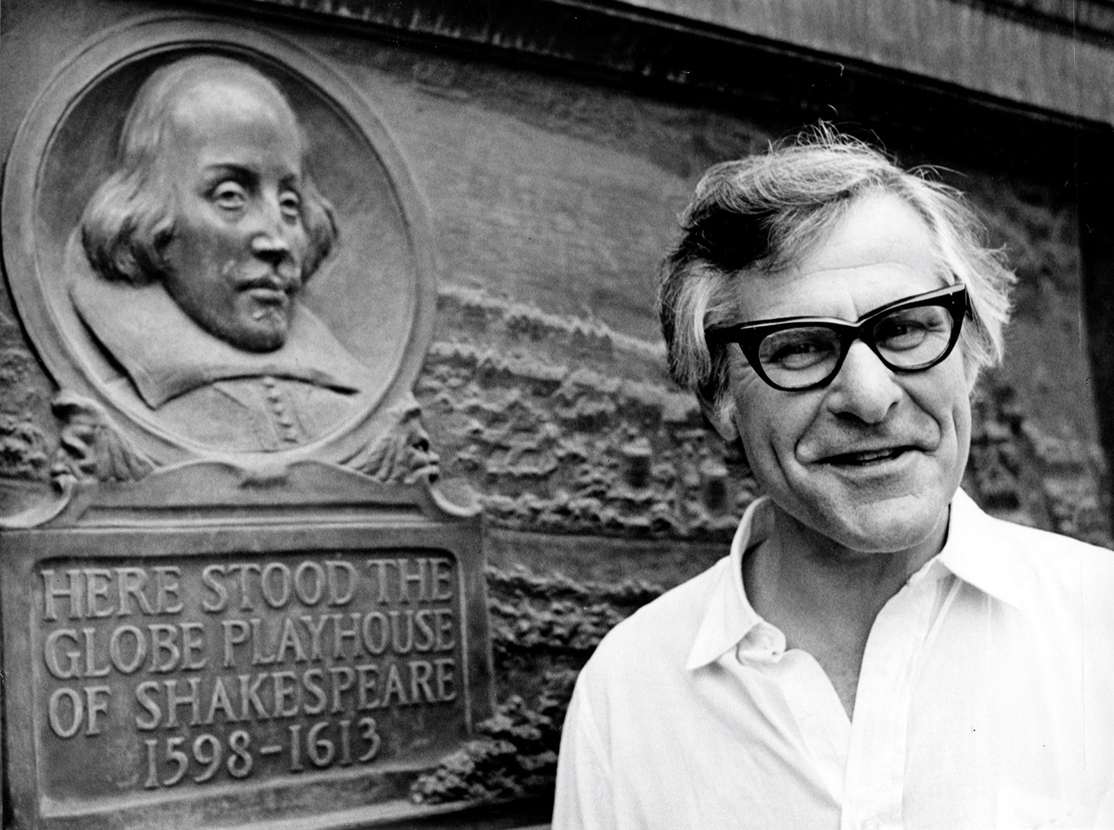 Sam Wanamaker Plaque Shakespeare's Globe A man wearing thick-framed glasses stands by a plaque marking the location of the original Globe Theatre.