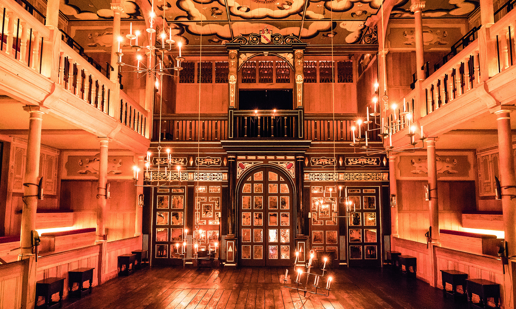 Sam Wanamaker Playhouse The candlelit interior of an indoor playhouse, with dark wooden panelling and a golden glow.