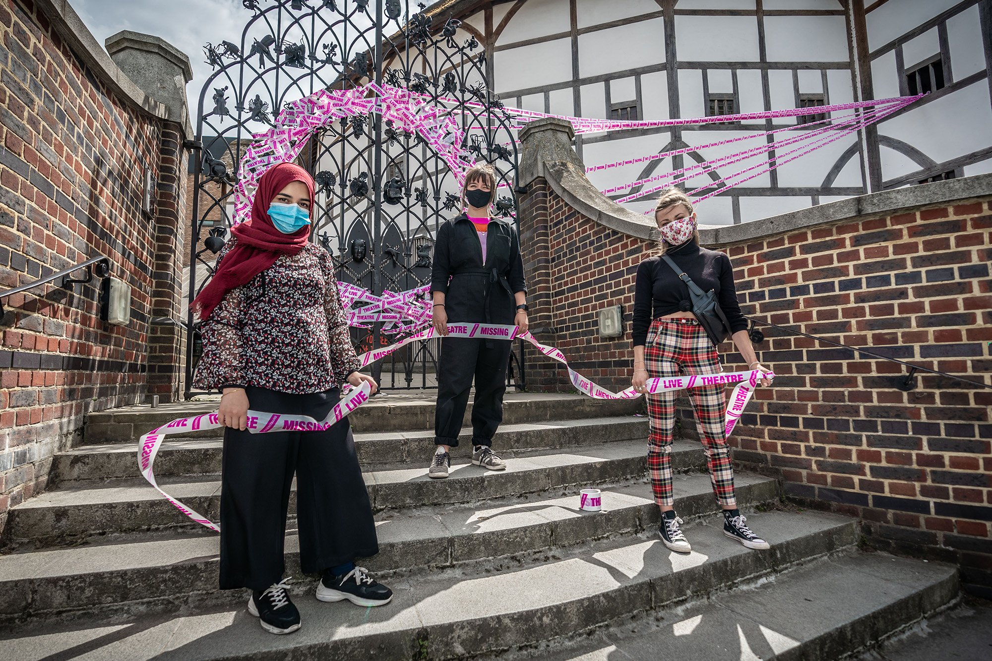 Scene Change Globe-21 flush Three women stand on a set of steps holding a length of white and pink type, which is formed in a circle on a set of iron gates.