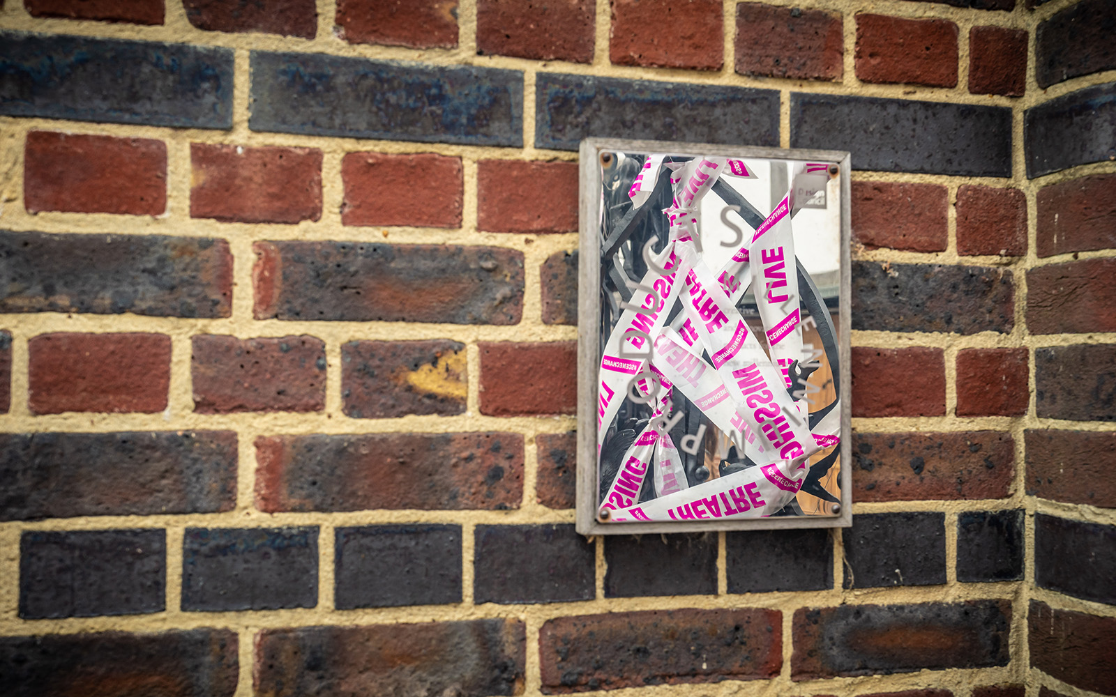 Scene Change Globe-47 A mirror on a brick wall reflects a criss-cross of white and pink tape.