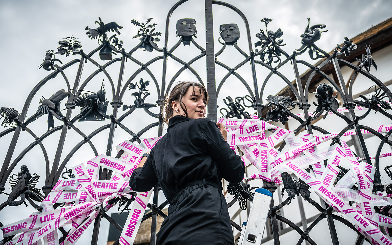 Scene Change Globe-69 A shot looking up at a woman standing before a set of iron gates, placing white and pink tape.