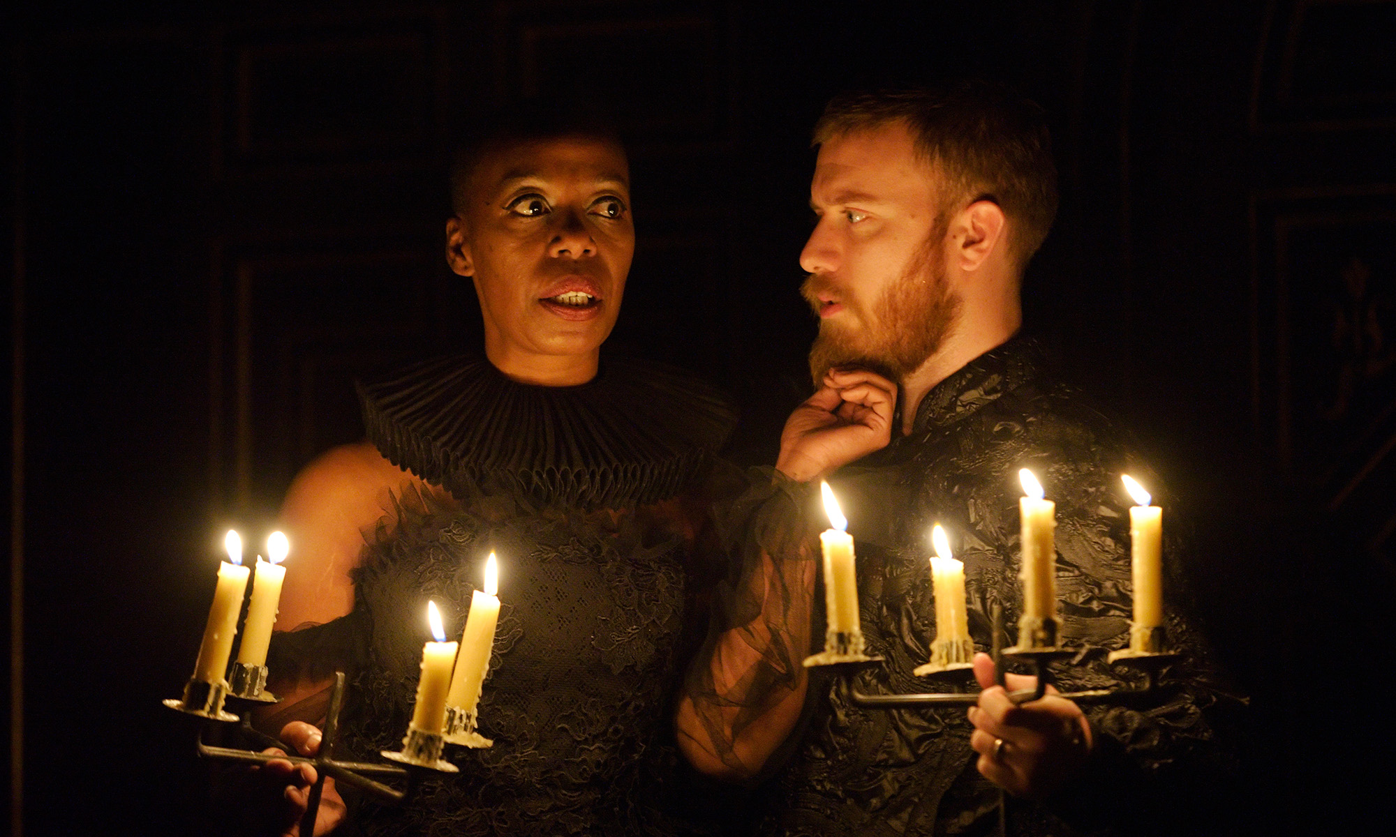 Tis Pity She's A Whore in the Sam Wanamaker Playhouse A woman and man stand in the glow of candlelight, the woman's hand touching the man's chin.