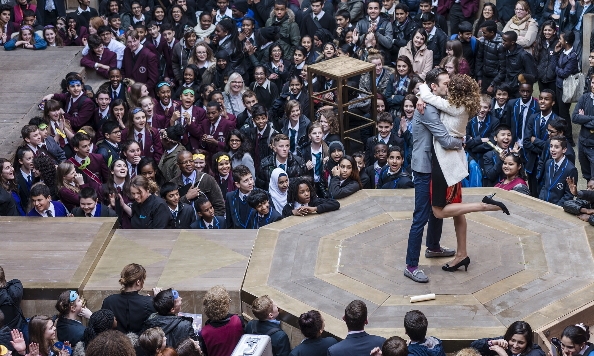Playing Shakespeare with Deutsche Bank Two actors kiss on stage, on a platform that extends into the audience in the yard