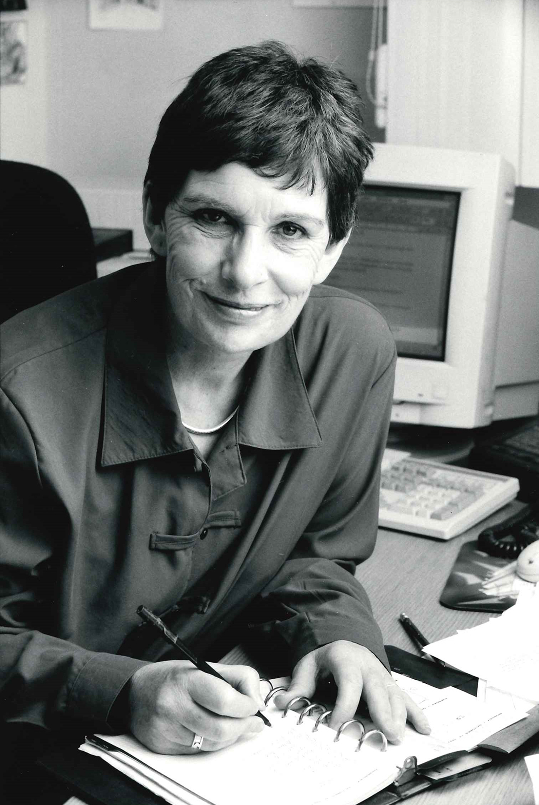 Globe Magazine Article Diana Devlin 1980s Portrait A black and white photograph of a woman smiling into the camera whilst writing in a notepad