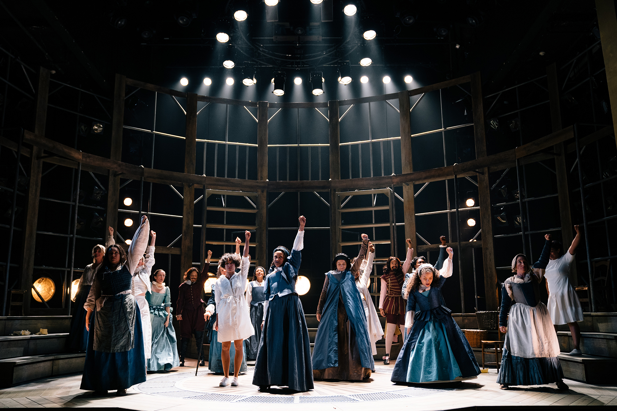 Emilia West End Shakespeare's Globe 2019 Helen Murray A group of actors stand on stage under spotlights, their left arms raised in the air into a fist.