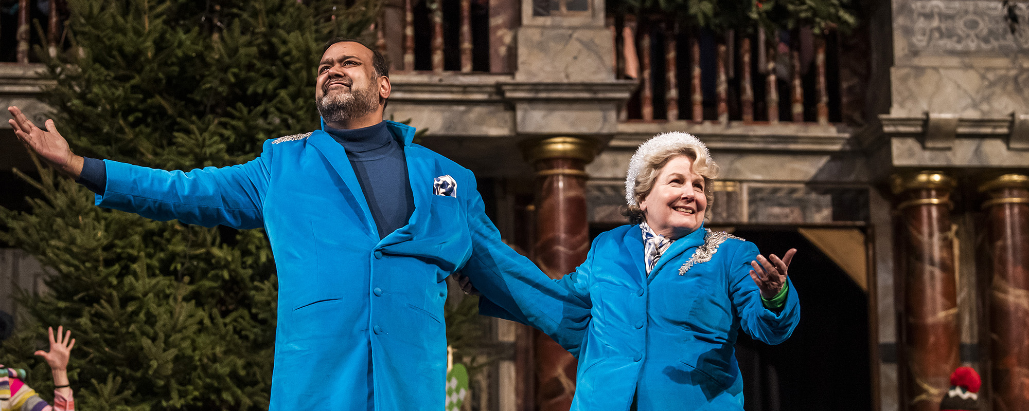 Christmas At The Snow Globe Competition Globe Advent 2 A male and female actor, wearing matching blue suits, stand onstage with their arms outstretched, smiling.