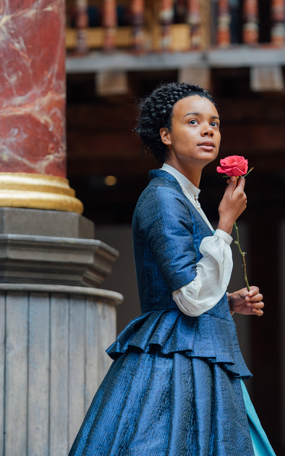 Emilia 1 Leah Harvey Shakespeare's Globe Helen Murray A woman wearing a blue Elizabethan corset dress holds a rose to her body.