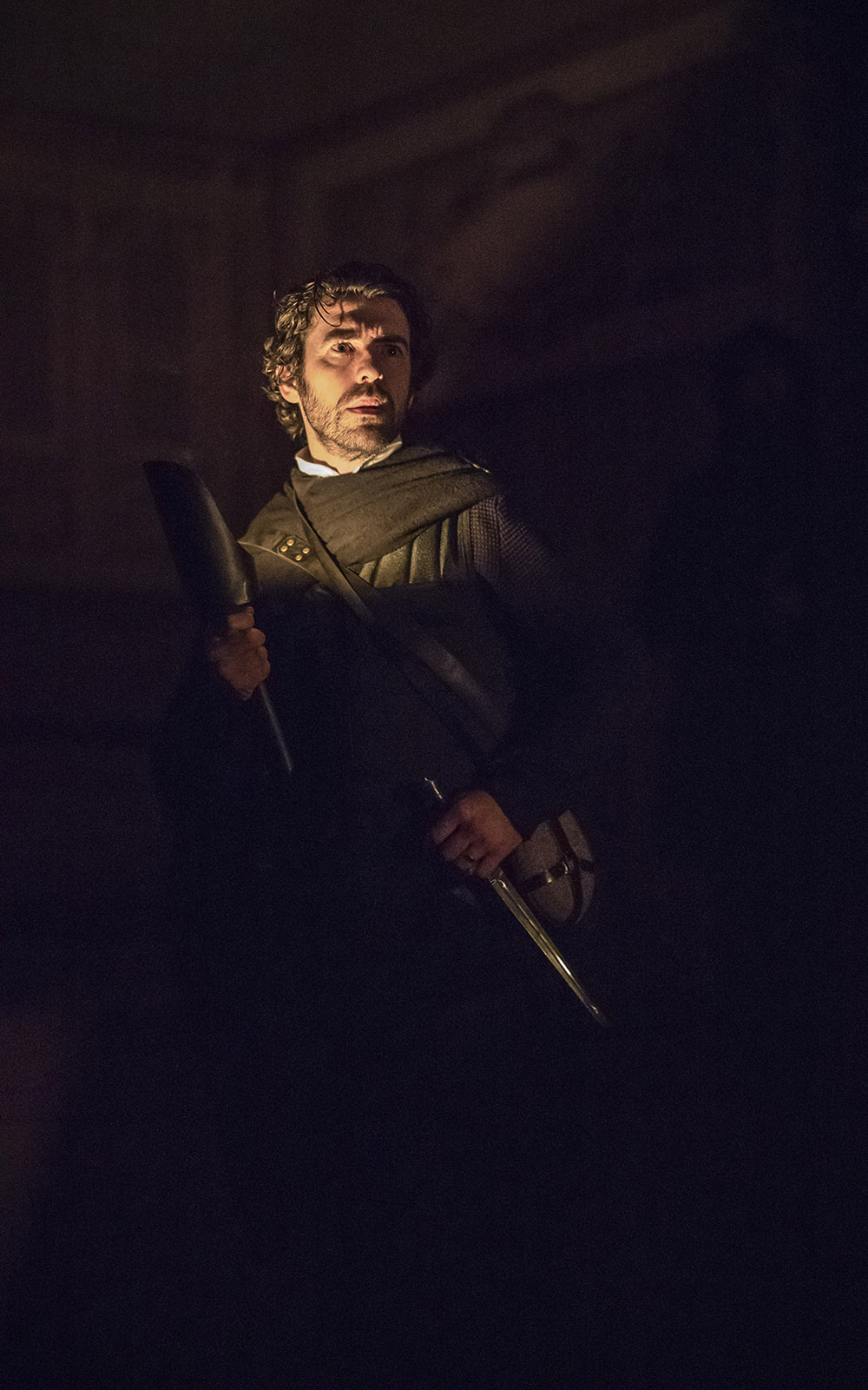MACBETH A fearful looking man stands holding two knives in the dark shadows of an indoor playhouse.