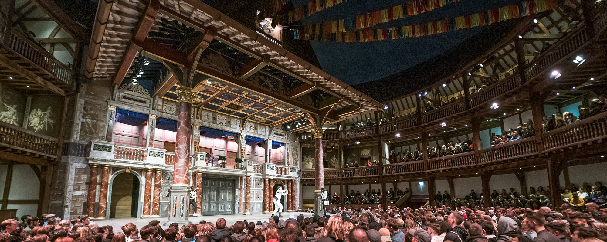 Giving Tuesday Shakespeare's Globe 2020 A full audiences watches an actor on stage at nighttime in a circular wooden theatre.