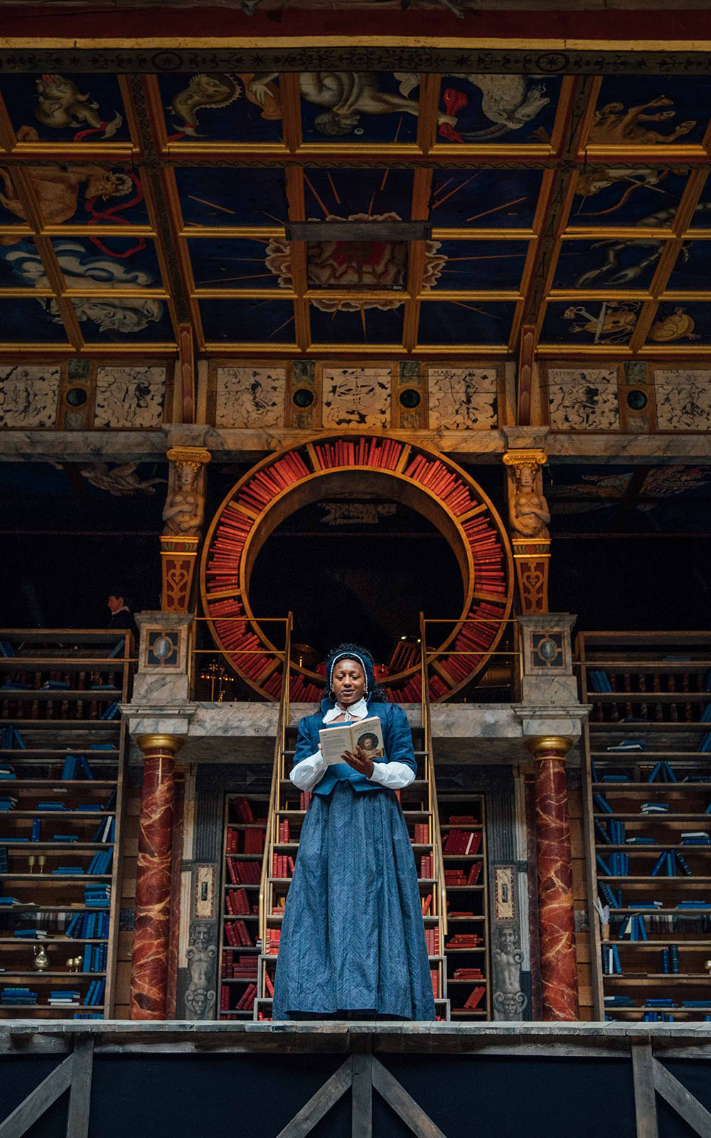 Blog Emilia Shakespeare's Globe A woman wearing a blue Elizabethan corset dress stands on stage, reading a book, with shelves of bookcases behind her.