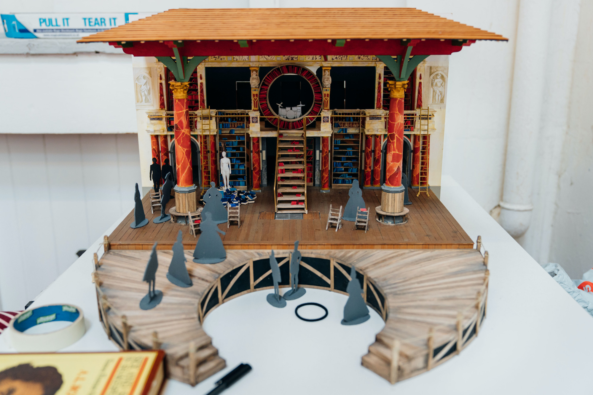 Emilia Model Box Costume Design Jo Scotcher Shakespeare’s Globe A mini set of a theatre which shows a wooden roof, wooden stage, steps, pillars and cardboard cut-out characters
