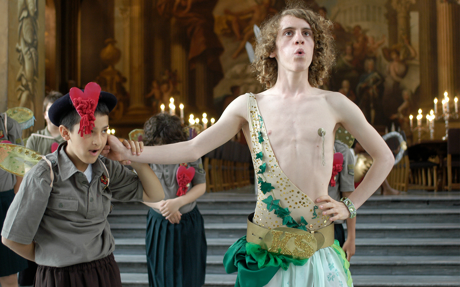 Galatea Such Stuff LGBTQ+ History Month Card An actor dressed in a gold leotard and green skirt has his hand around the neck of a young actor
