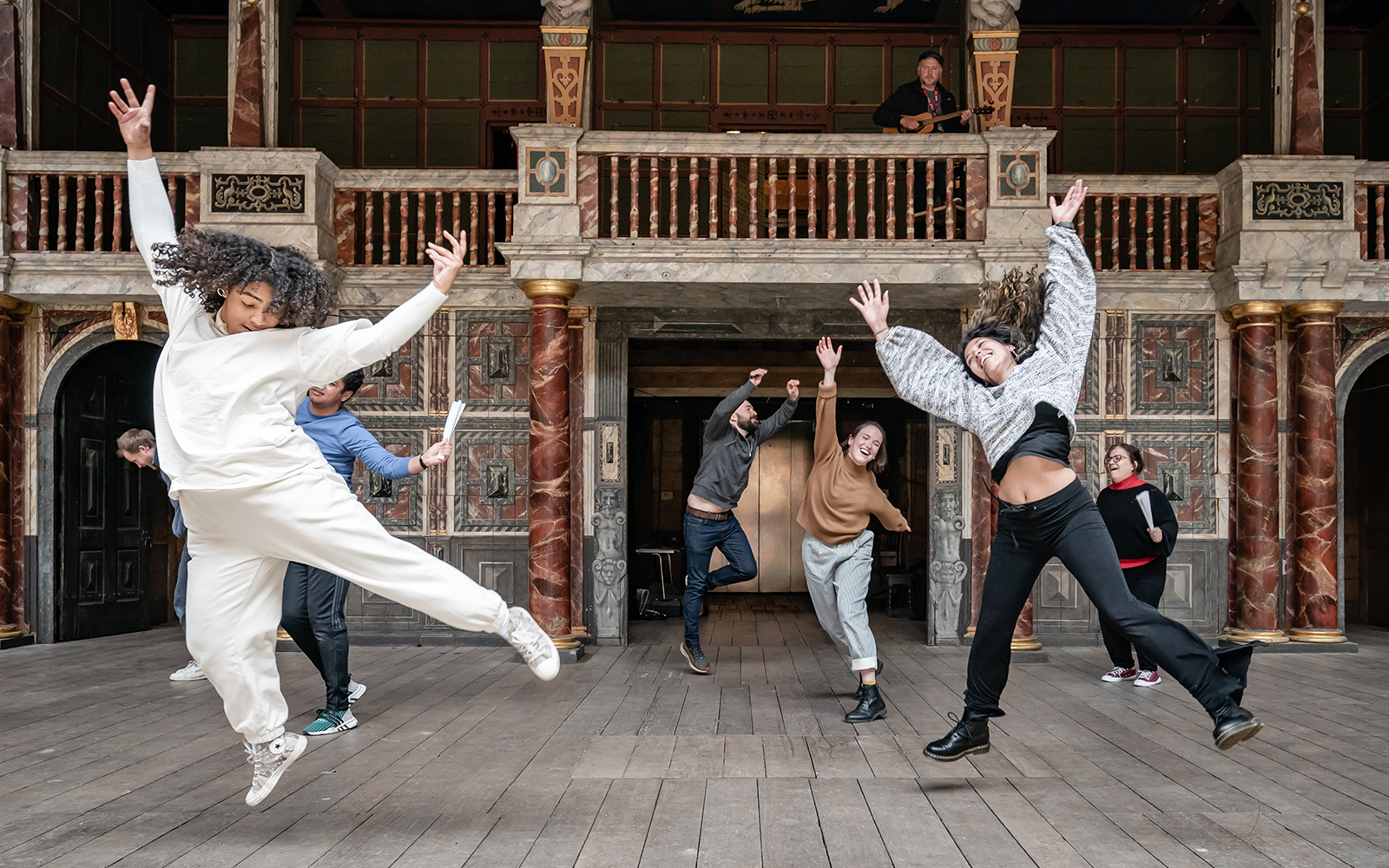 Globe on Tour 2021 Rehearsal Image As You Like It On an outdoor, wooden stage, a group of actors dance and jump in the air