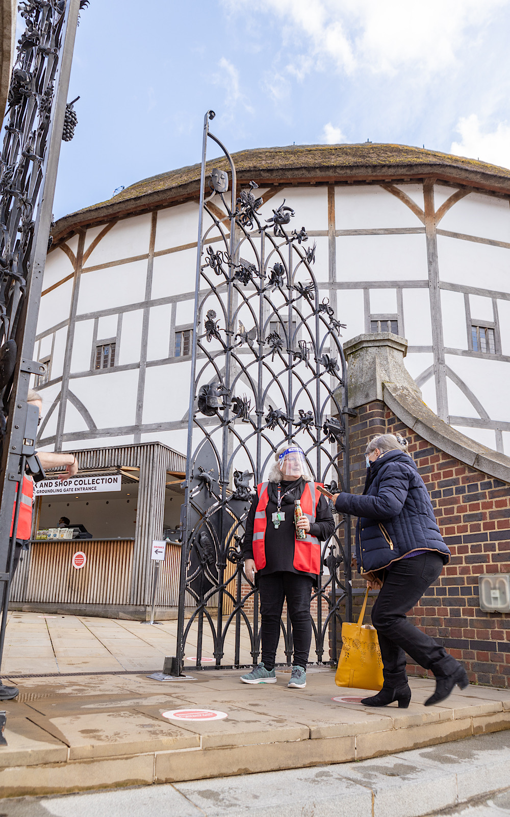 Covid Coronavirus Shakespeare’s Globe Pete Le May 2021 An audience member walks up stone steps and through a black, metal gate where a member of staff in a visor greets them