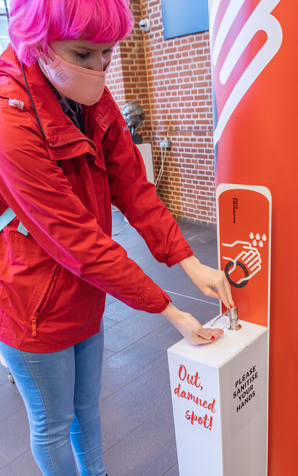 Covid Coronavirus Shakespeare’s Globe Pete Le May 2021 An audience member in a red jacket leans down to sanitise her hands at a sanitiser station