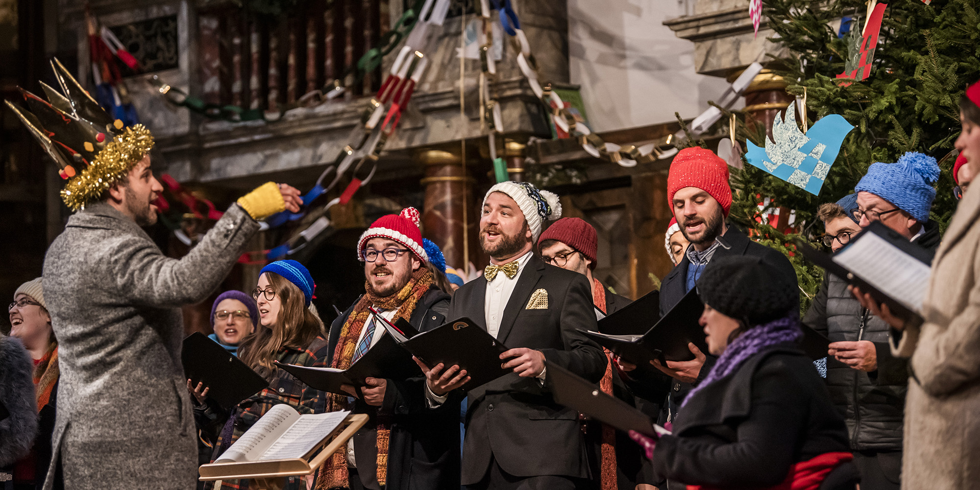 Christmas At The Snow Globe-Shakespeare's Globe 2021 A group of choral singers wearing festive gear and Santa hats, stand on the Globe Theatre stage in front of a decorated Christmas tree, singing.