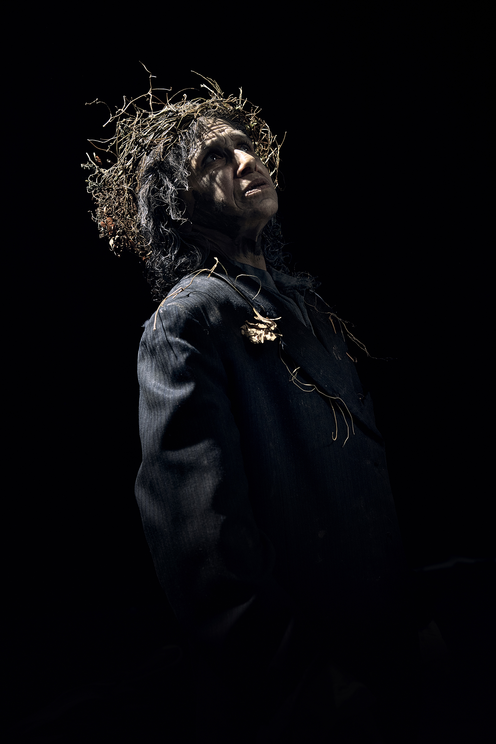 Shakespeares Globe_Kathryn Hunter_King Lear_Photographer Kate Bones_2022 An actor wearing a wreath crown of branches and leaves looks up, their face cast in shadow.