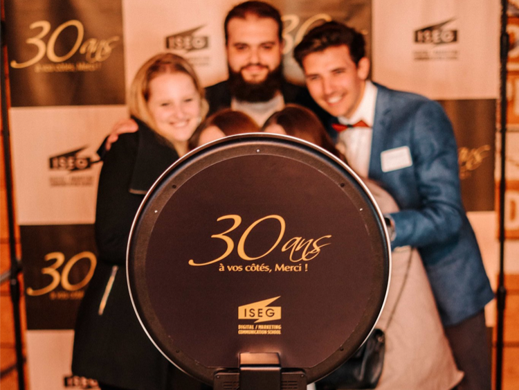 Un groupe d'amis se prend en photo avec le photomaton de Josepho devant un photocall aux couleurs et logo de l'école ISEG. La borne photo de Josepho est personnalisé avec le logo de l'ISEG.