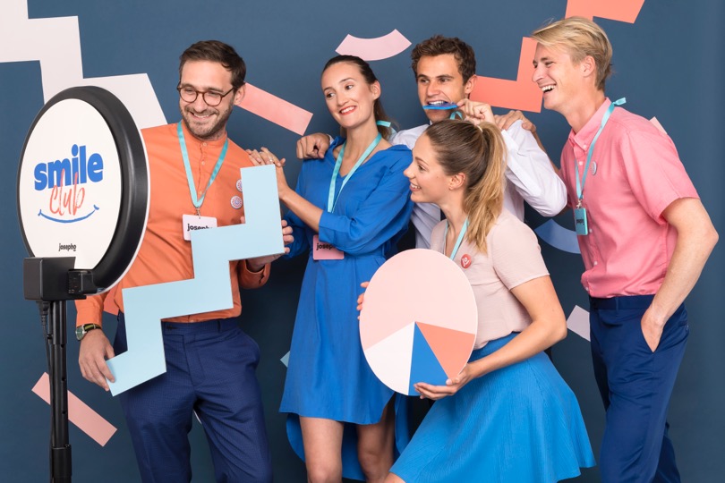 Un groupe de 5 collègues sourit et se prend en photo devant le photobooth de Josepho qui est personnalisé avec le slogan "Smile Club" de Josepho.