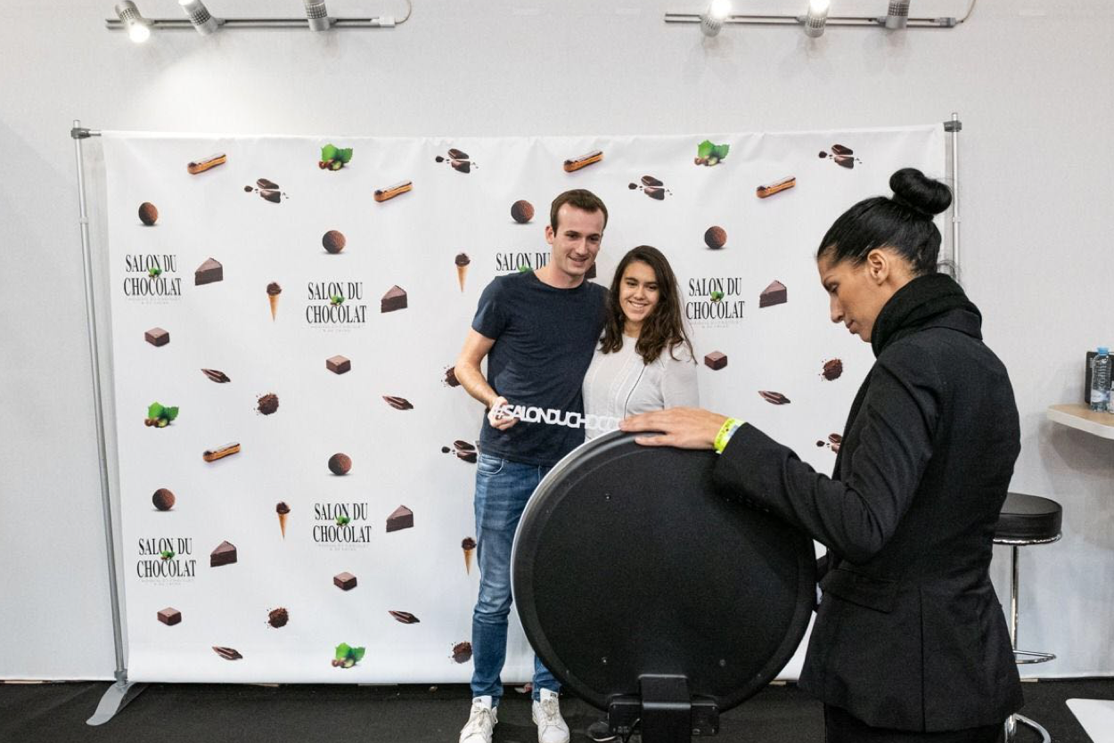 Sur la Salon du Chocolat, une hôtesse d'accueil prend en photo un couple avec le photobooth de Josepho devant un photocall aux couleurs du salon.