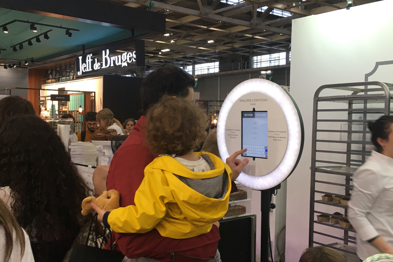 Sur le Salon du Chocolat, un père avec son enfant des les bras pianote sur l'écran du photobooth de Josepho qui est personnalisé avec le logo Philippe Conticini.