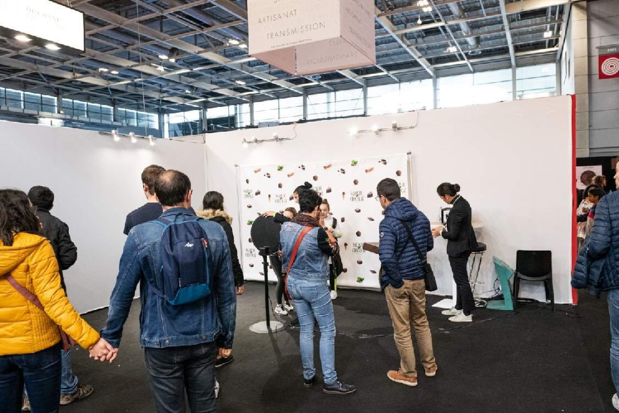 Sur le Salon du Chocolat, le photobooth de Josepho est installé face à un photocall aux couleurs du salon. Des enfants sont en train de se prendre en photo avec le photobooth et une hôtesse les assiste.