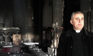 Father Benham stands amongst the ruins of his church; a smashed altar behind him is a tribute to the force of destruction that moved through it.