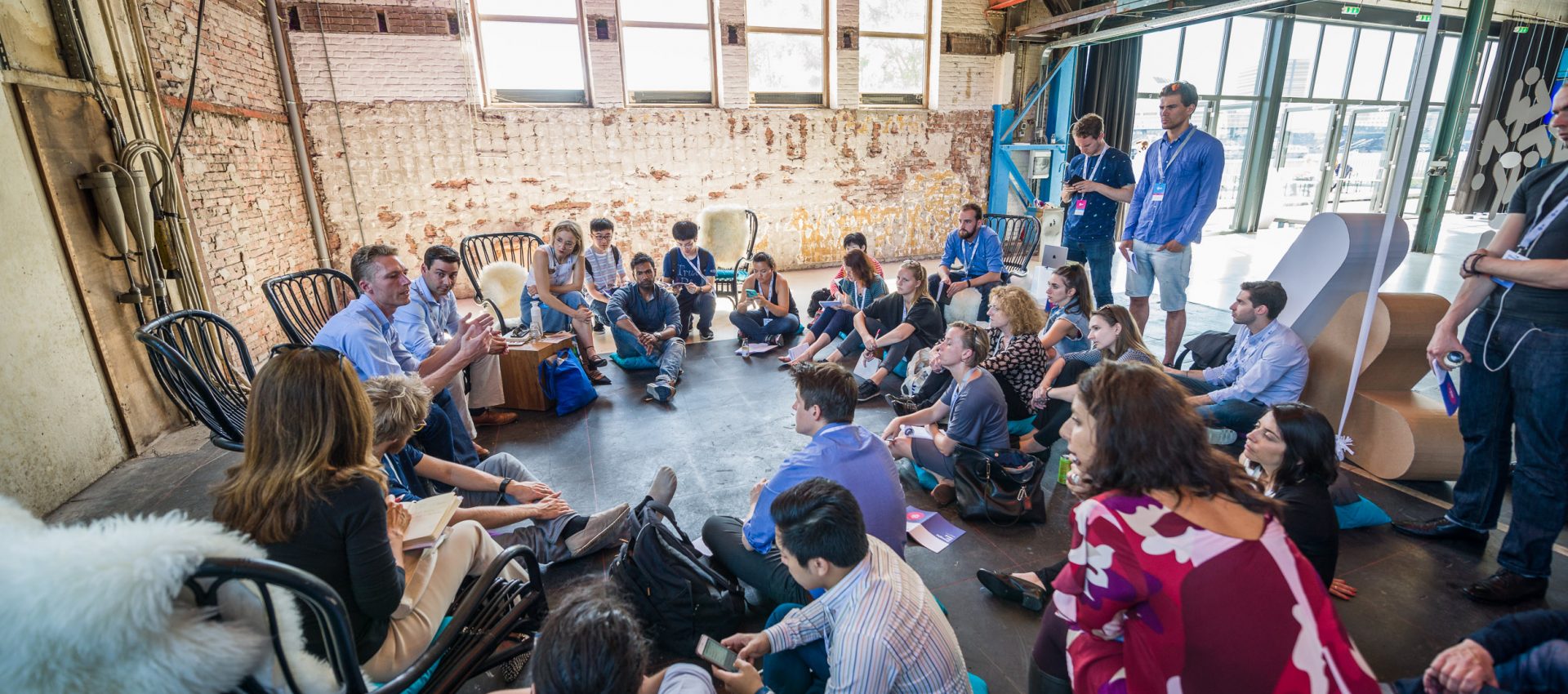 TFF Challenge participants sitting in a circle in a factory-like room, they are engaged in an active discussion. Three men are standing and watching / listening the conversation.