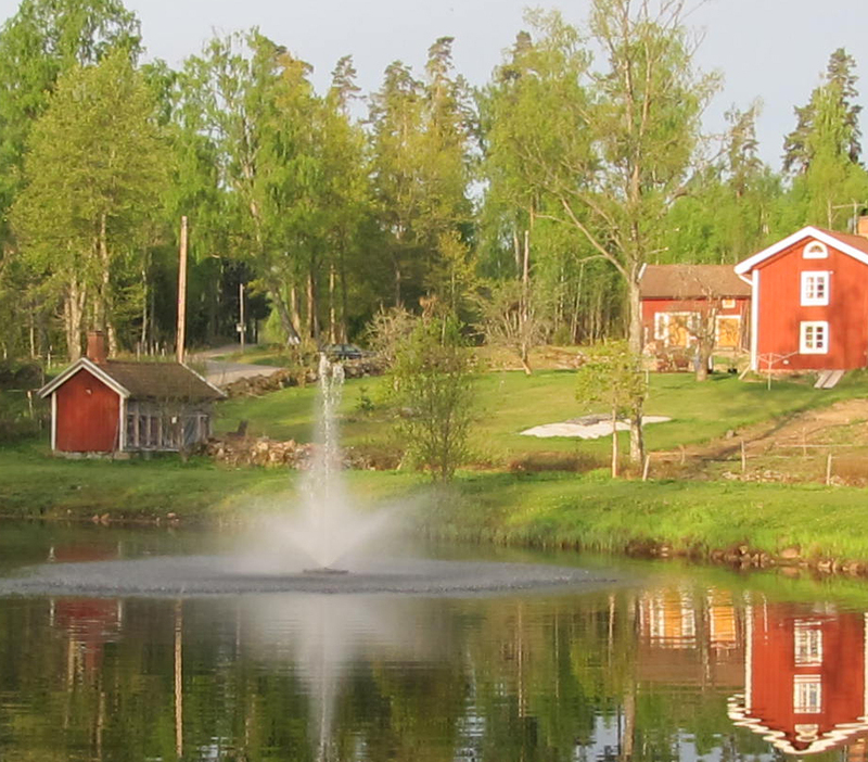 Kasco flytande fontän i damm framför rött hus med vita knutar på landet. 