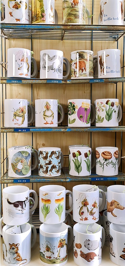 Foto på en hylla i butiken. Hyllan är fyllt med muggar, muggarnas motiv är med kor, hundar, melonpingviner, taxar, kaniner.