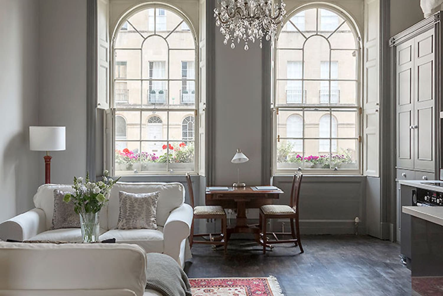 The open-plan living space of an apartment including table and chairs for two, sofas, a chandelier and two large windows.