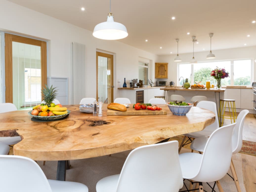 A kitchen/dining room with breakfast bar, a wooden table and white chairs, flowers on the counter and fruit, veg and bread on the table. 
