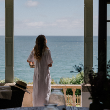 A woman in a long white dress looks out from a doorway across the sea